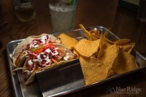 Close up of two Tacos in a corn shell in taco holders, garnished with cabbage and sauce. Chips on the side with a small container of red Salsa