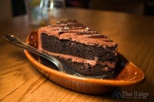 Close up photo of a slice of chocolate cake from Coyote Kitchen.  The cake is two layers with chocolate icing between the layers and on top with two spoons on the brown plate next to the cake.
