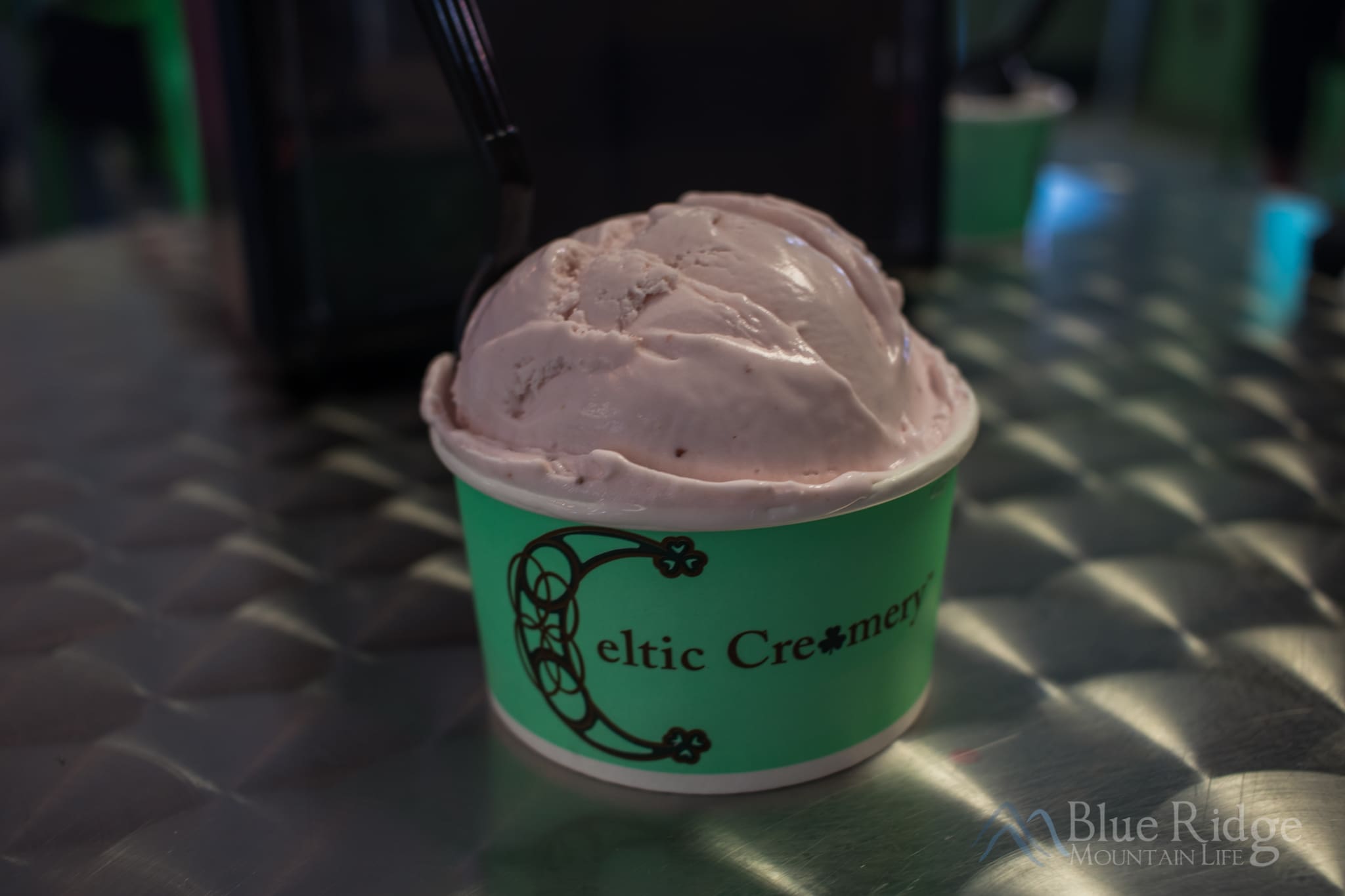 Strawberry Ice Cream At Celtic Creamery Photo of a single huge scoop of pink strawberry ice cream on a metal table. The Celtic Creamery logo is on the green paper cup.
