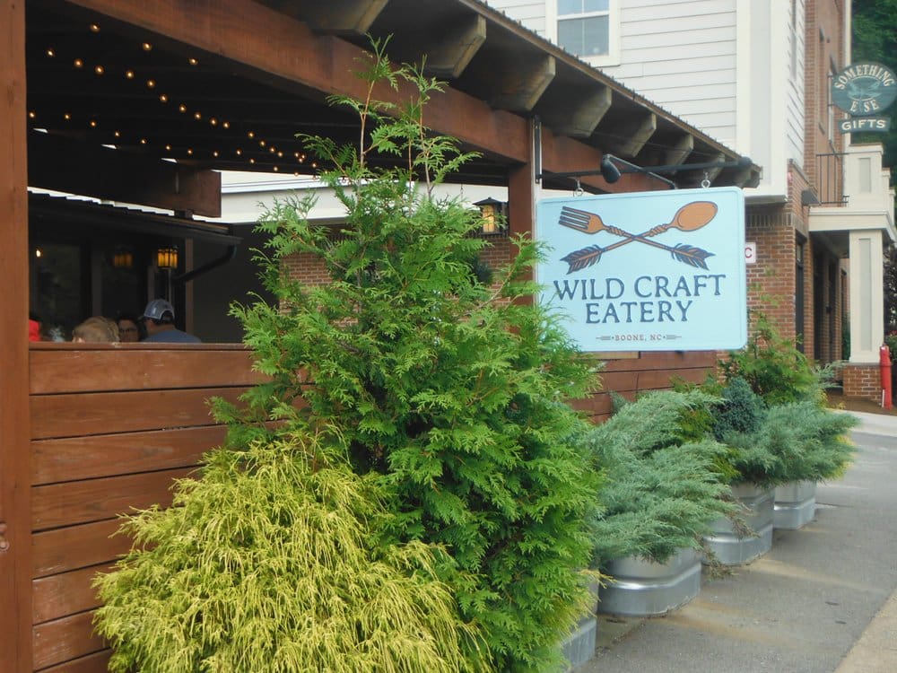 Wild Craft Eatery Boone NC Front of Wild Craft Eatery Boone NC with their covered outdoor seating area, surrounded by green shrub. Their sign is hanging visibly on the front.