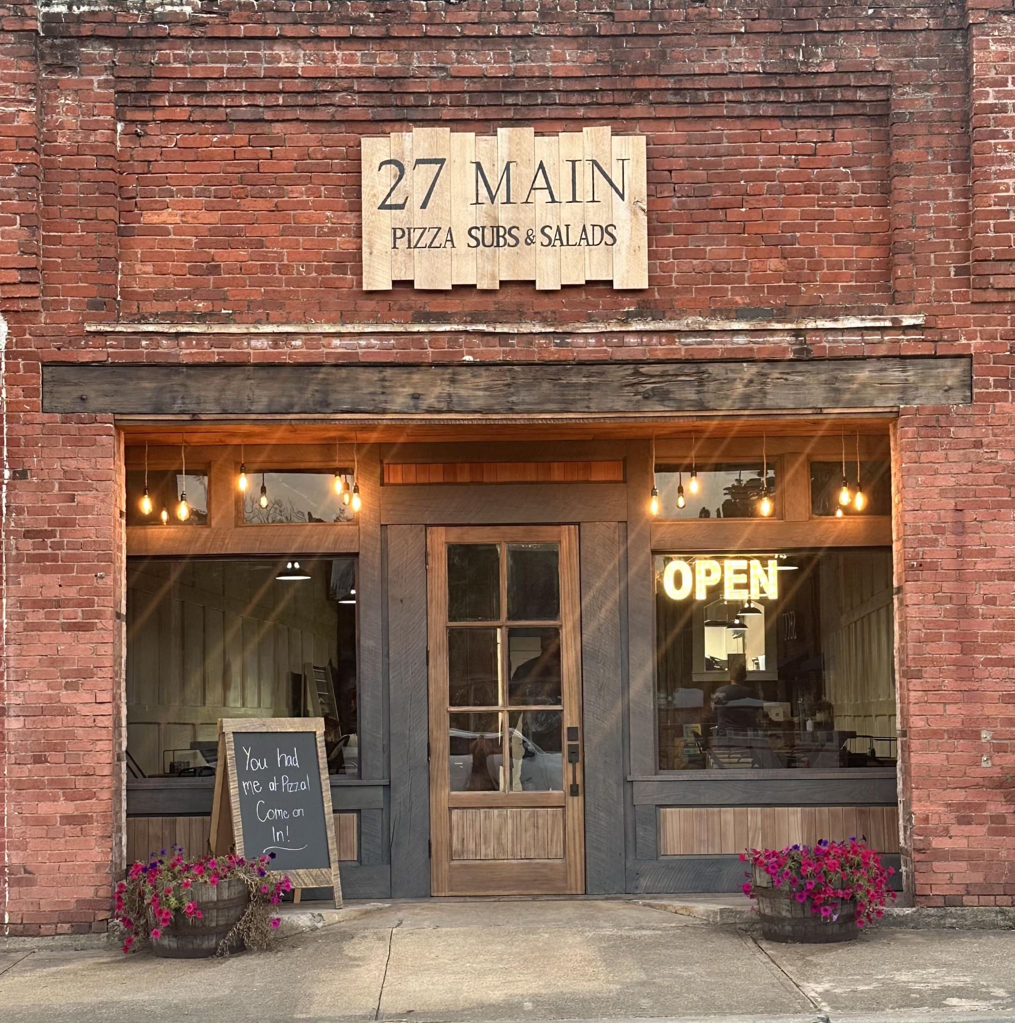 27 Main Restaurant Entrance Front of 27 Main showing its brick front, wooden entry door, and warm lighting. The 27 main sign is made of wood boards, giving it a rustic look and feel.