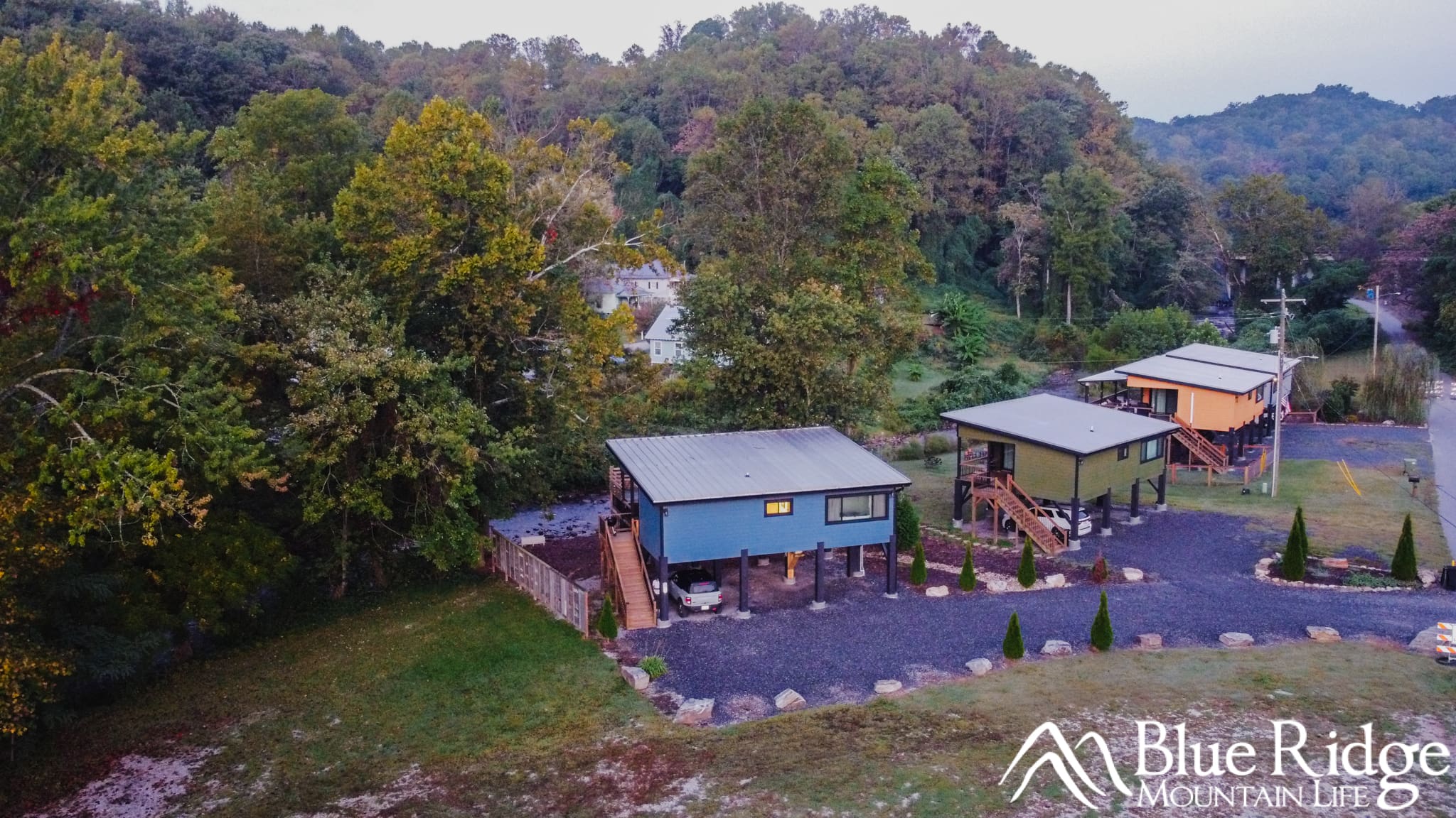 Old Fort Rental Cabin Drone photo of our blue rental cabin on stilts next to the creek and surrounded by green trees. There is a gravel driveway in the front, with stairs on the side leading up to the main livin floor. A bronco sport is parked in the parking area under the cabin.
