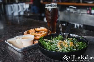 Kale Salad on a black plate.  Beside it are the the hot soft pretzels with beer cheese in a plastic cup.  Behind all of this is a large glass of dark amber beer.