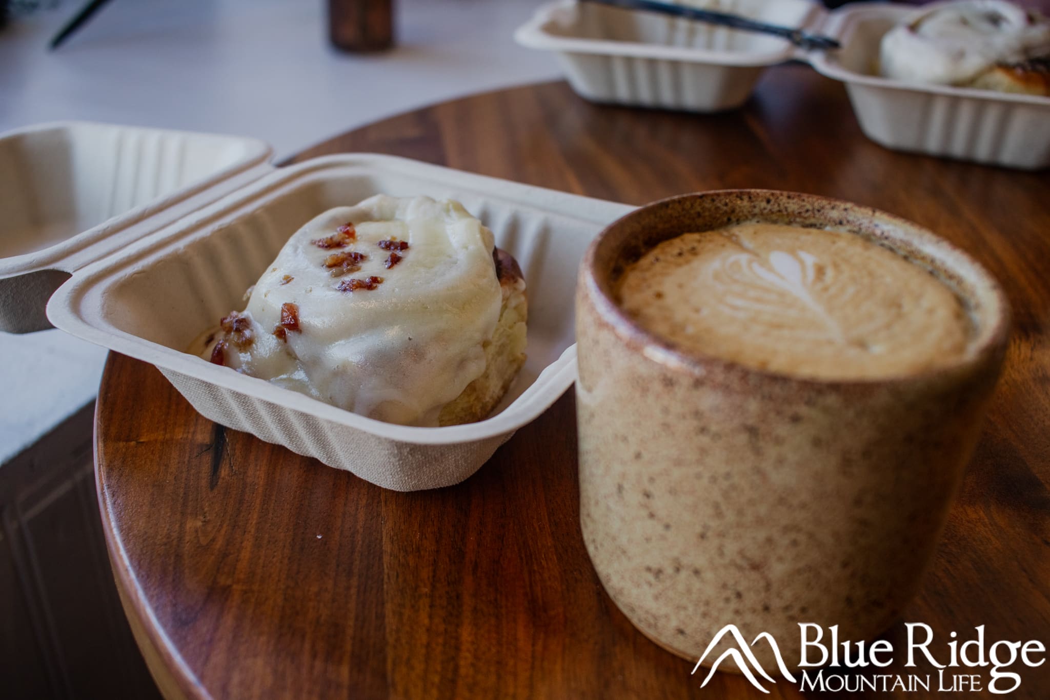 Gogos Cinnamon Rolls Old Fort NC Maple Bacon Cinnamon Bun with white creamy icing on top, next to a Cappachino coffee in a brown speckled mug