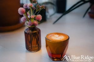 Photo of a latte coffee in a semi-transparent brown glass, next to a small pink flower arrangement.  The coffee has a flower on top in the foam.