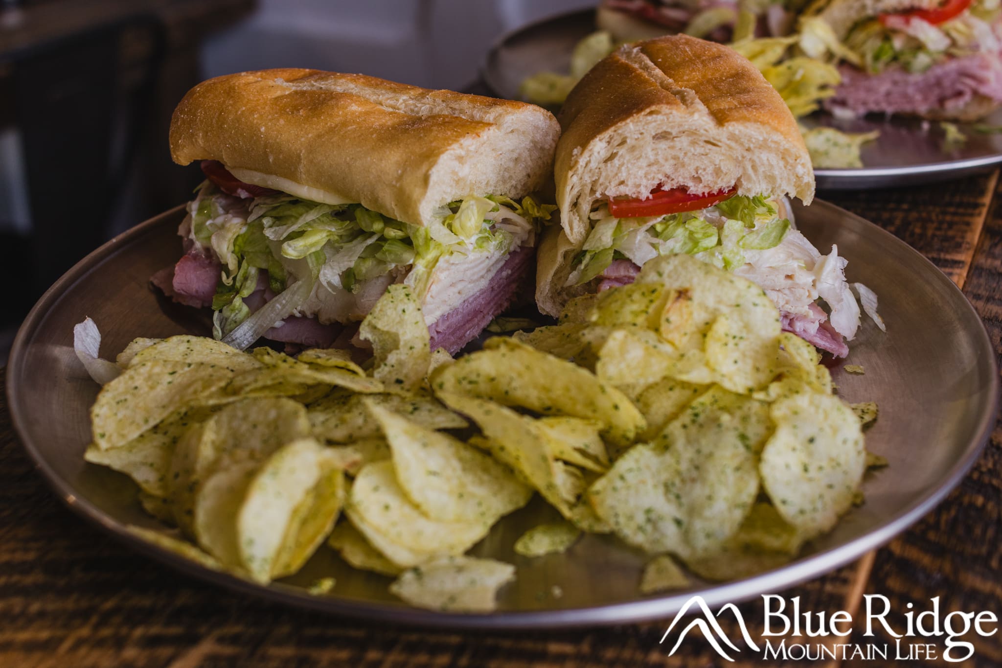 Cold Cut sandwich with a sub roll, ham, turkey, lettuce and tomato served with chips on a metal plate. The sandwich is cut in half.