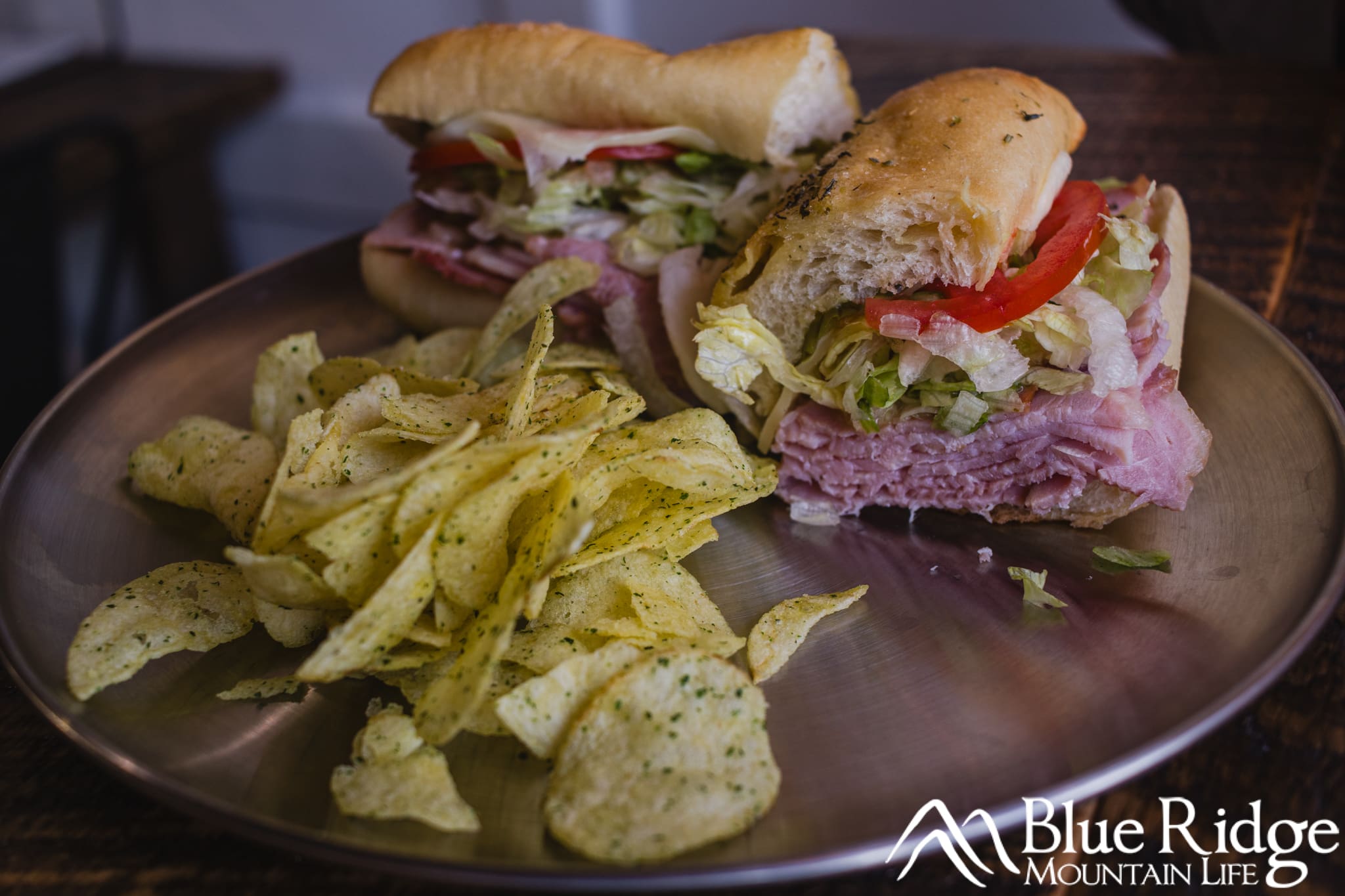 Italian sandwich with a sub roll, ham, lettuce and tomato served with chips on a metal plate. The sandwich is cut in half.