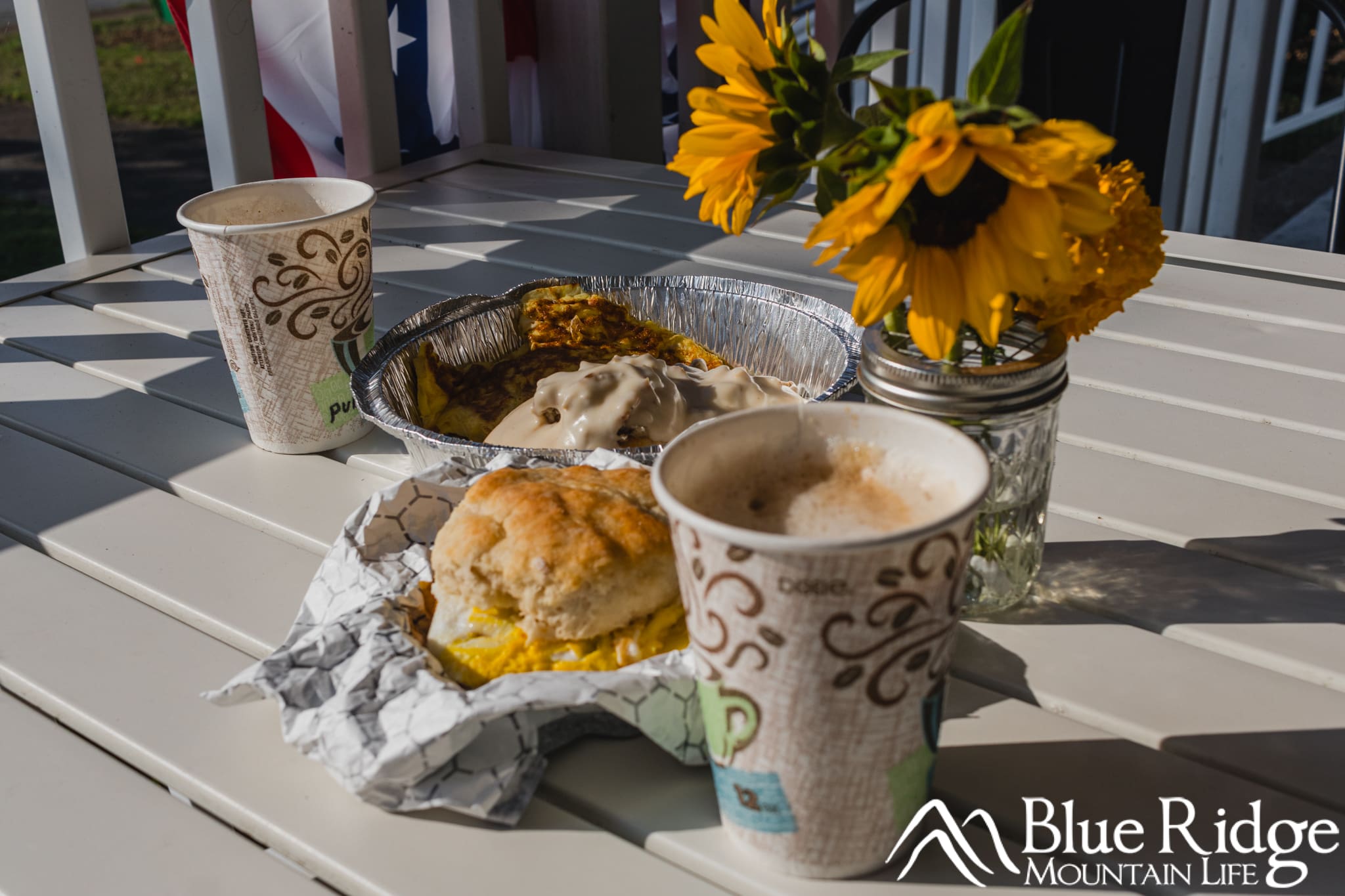 Breakfast at the Davis Country Store and Cafe Photo of the biscuits in gravy in a metal togo container, with a bacon, egg and cheese biscuit in front of it and a coffee to the side. The food is arranged around a yellow flower arrangement on the table.