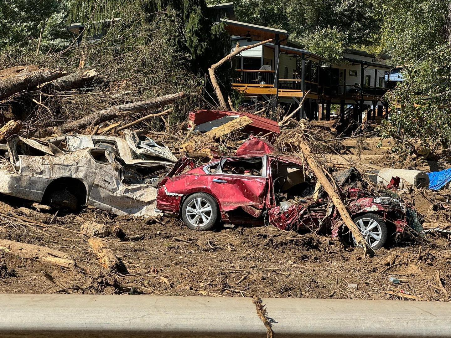 Hurricane Helene damage to Old Fort, NC Flooding damage to Old Fort, NC. Photos shows two cars with significant damage, all bent with parts missing. Surrounding the cars is debris from trees and mud all over the ground. A damaged rental home is in the background.
