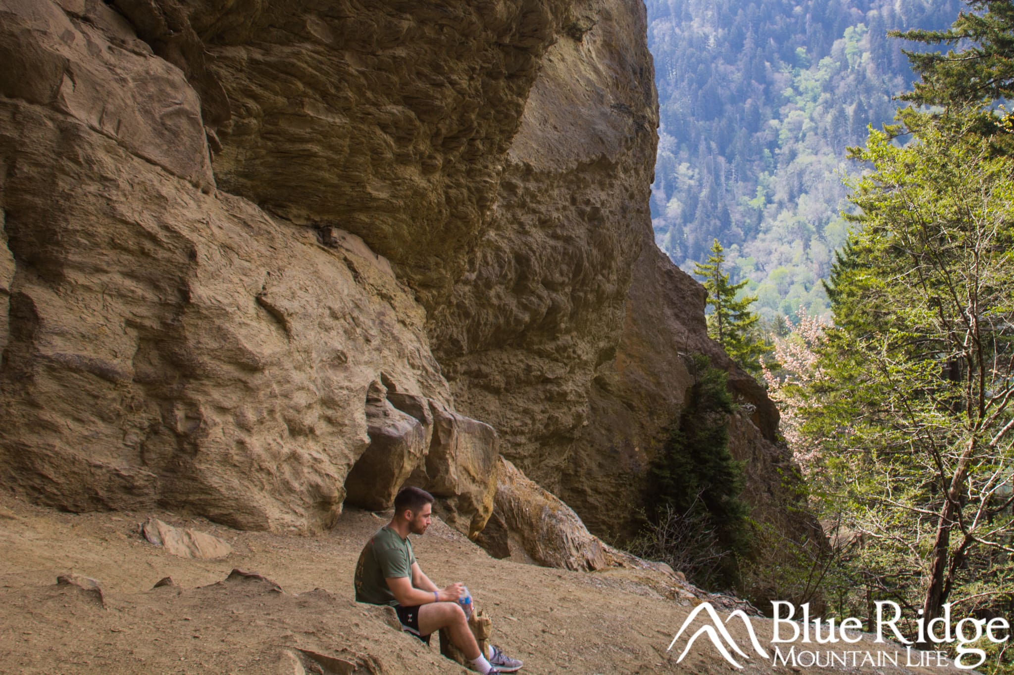 Alum Cave Alum Cave - Photo of an well built young male sitting on the ground, with the large brown rocks of a cave behind him. To his front is a beautiful mountian view.