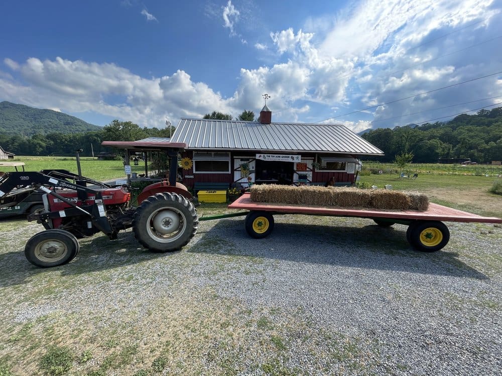 10 Acre Garden Canton NC Photo of a tractor and trailer in front of a farm building with blue skies and green fields in the background.