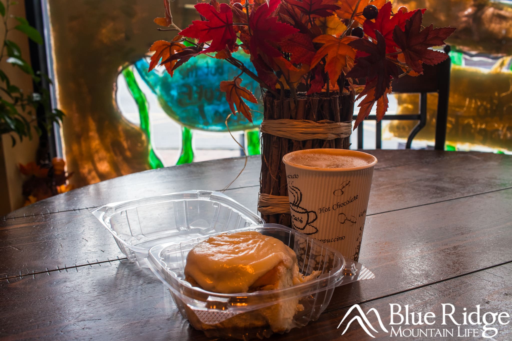 Coffee and a Cinnamon Bun at Ingenious Coffee Hot Latte in a paper cup next to a Cinnamon Bun. Both placed on a table, alongside a fall themed flower.