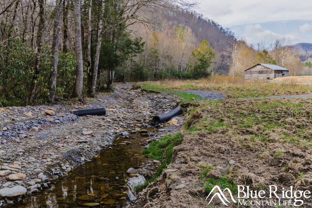 Photo of Cataloochee Valley After Hurricane Helene. Photo shows washed out road on the left, with drainage piping exposed. Right side shows Caldwell Barn in the distance, with a temporary gravel road for access by park staff