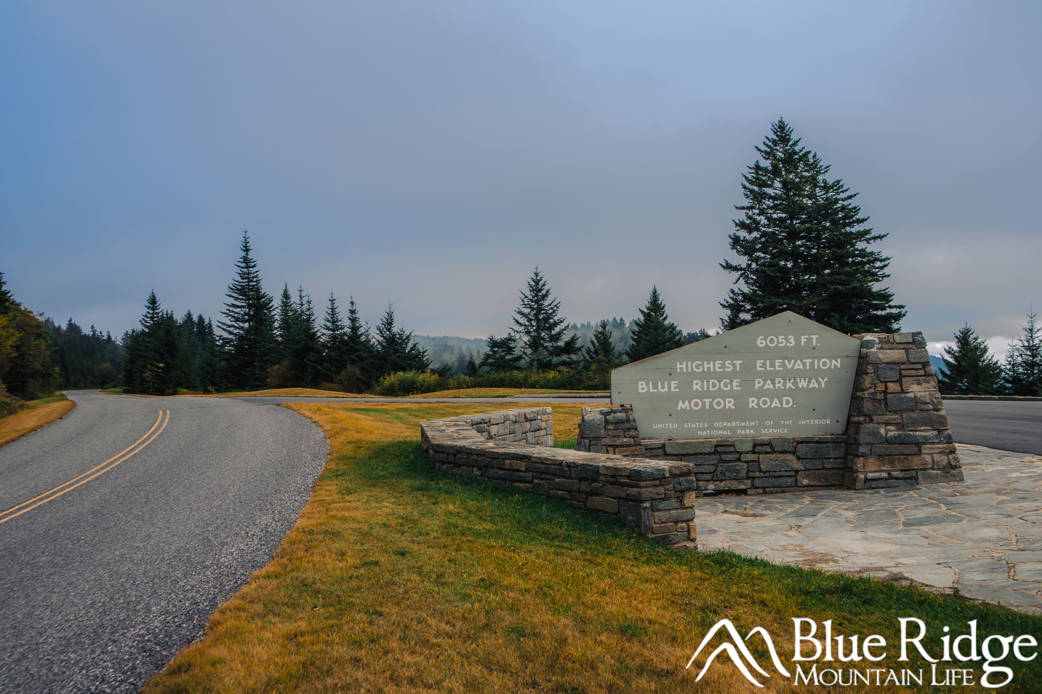 Highest Point on the Blue Ridge Parkway Photo of the Highest Point on the Blue Ridge Parkway, showing the parkway curving to the left and an overlook on the right with a large sign saying 6053 feet, Highest Elevation, Blue Ridge Parkway Motor Road. In the background are distant evergreen trees and mountains.