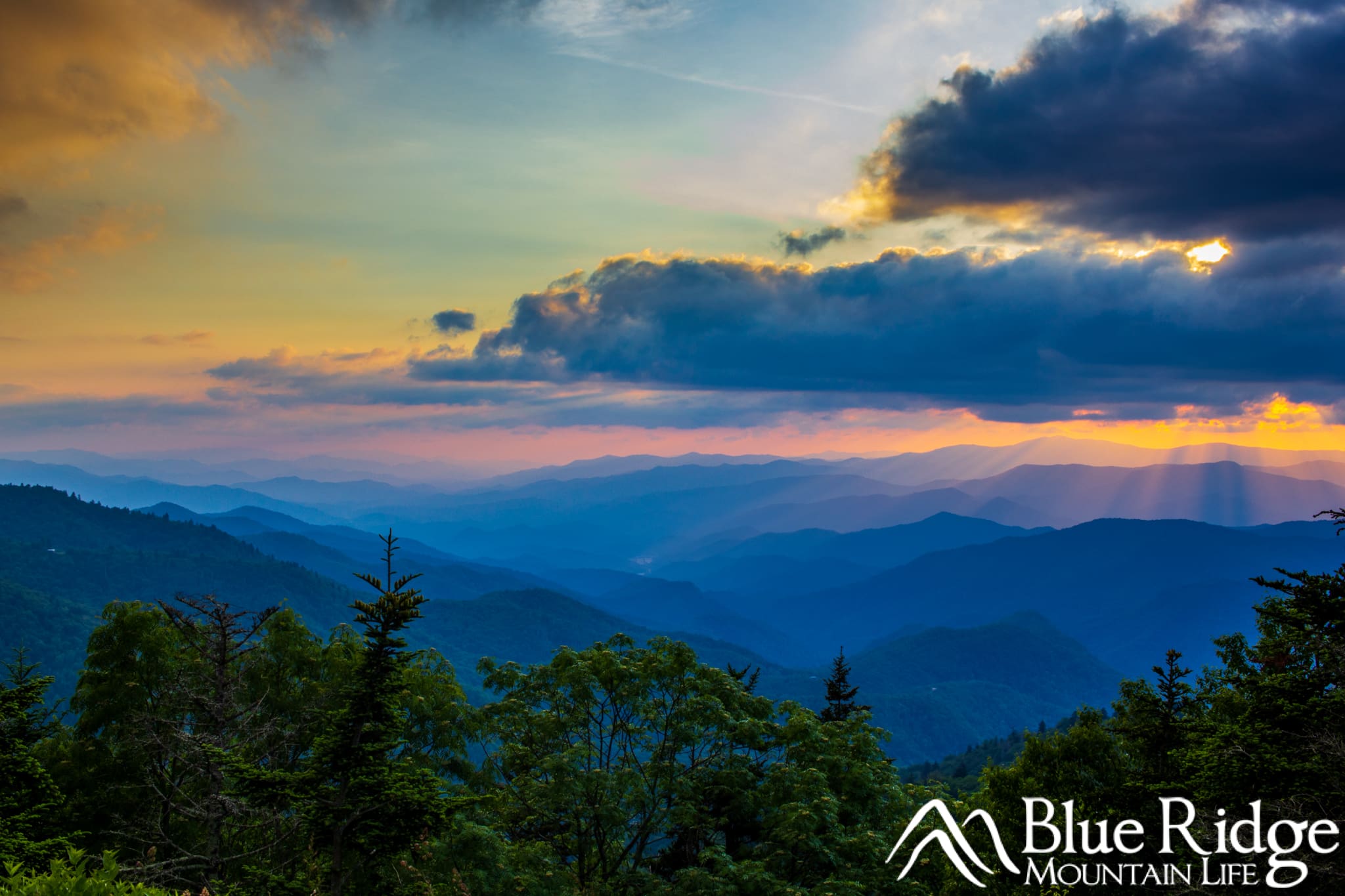 Waterrock Knob near Waynesville NC Sunset photo at Waterrock Knob showing distant blue colored mountains, with dark blue clouds in the sky. Orange light from the setting sun hovers between the mountains and clouds with sunrays coming down to the mountains.