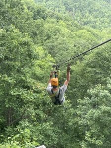 Larry zipping down a zipling with the forest below him and the mountains in the distance.